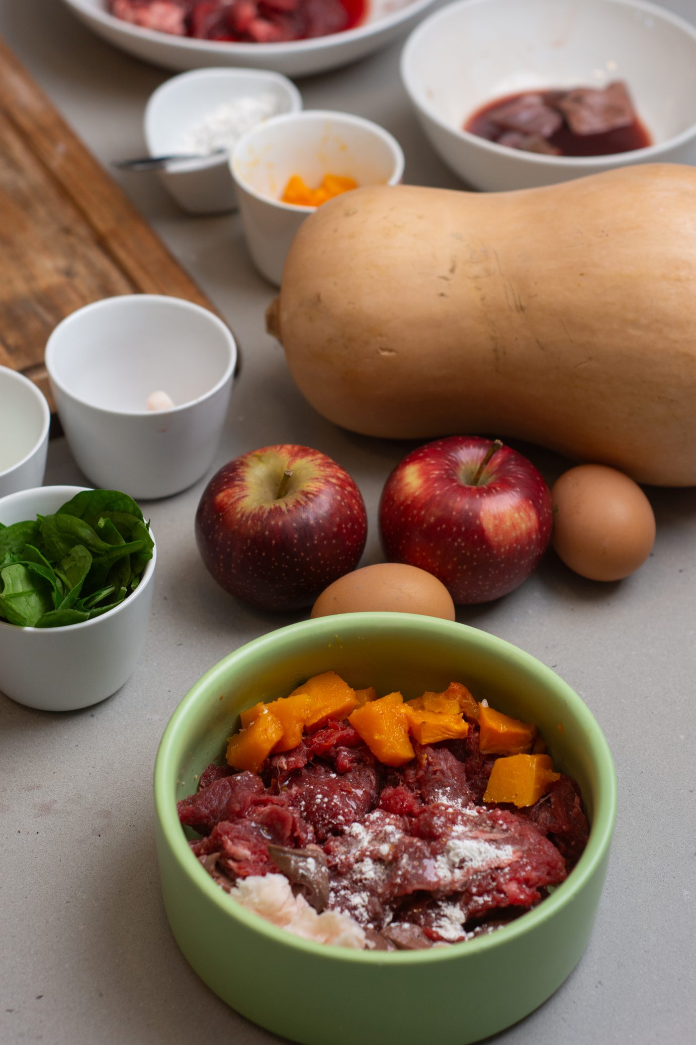 A balanced French Bulldog meal coming together in a normal kitchen: green bowl with beef and pumpkin, butternut squash, apples, spinach and eggs on the counter